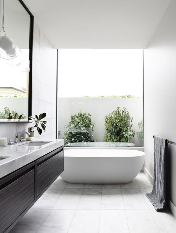 a light-filled contemporary bathroom with a black floating vanity, white marble tiles, a large tub and a glazed wall