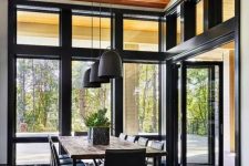 a lovely and simple contemporary dining room with a view of the forest, a simple stained table and black chairs, black pendant lamps