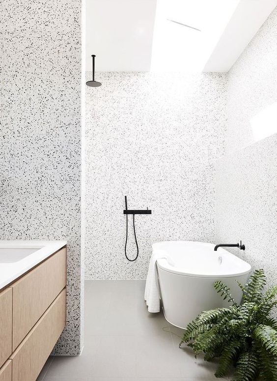 a lovely contemporary bathroom with terrazzo walls, grey tiles, a blonde wood vanity and an oval tub, black fixtures