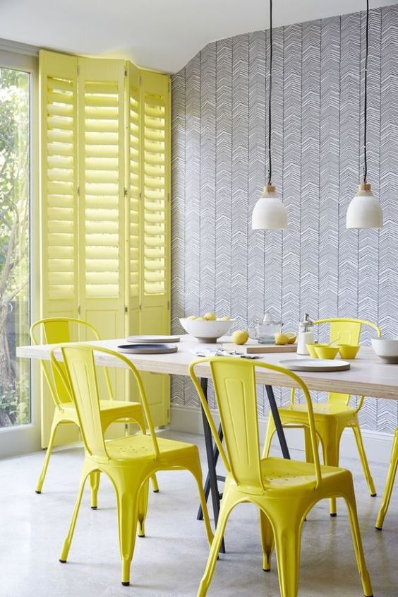 a lovely dining room with a printed accent wall, a stained table and yellow metal chairs, yellow shutters and pendant lamps
