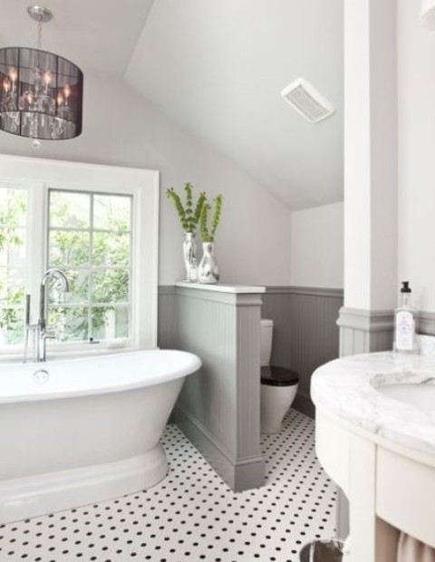 a lovely farmhouse bathroom with a tile floor, grey paneling on the walls, a vintage tub and a chic chandelier plus a half wall that separates the space