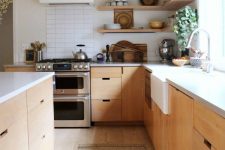a modern blonde wood kitchen with no hardware cabinets, white stone countertops, open corner shelves, a white tile backsplash