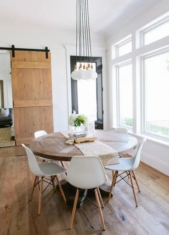a modern rustic dining room with a round table, white chairs, a cluster of pendant lamps and greenery plus lots of natural light