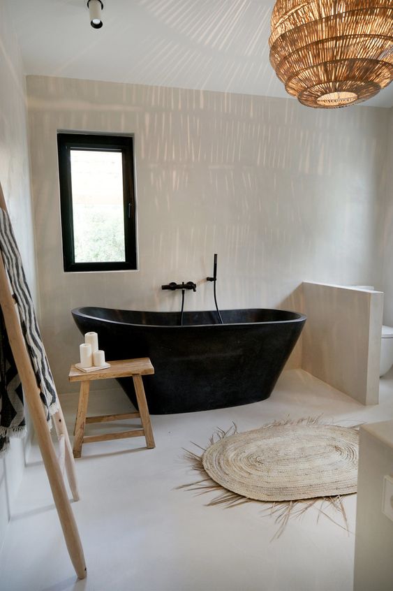 a modern tropical bathroom done in warm neutrals, with a catchy black bathtub and a half wall that separates the toilet from the rest of the space