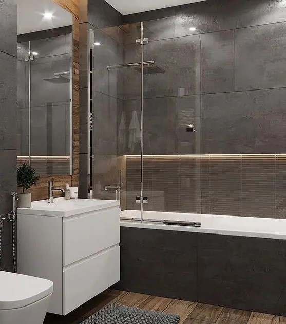 a moody contemporary bathroom clad with dark stone tiles, with built in lights, a wooden floor, white appliances and neutral fixtures