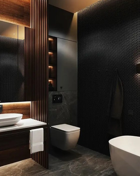 a moody contemporary bathroom with a black penny tile wall, a grey marble floor, a built in vanity, a wooden beam screen and a lit up mirror