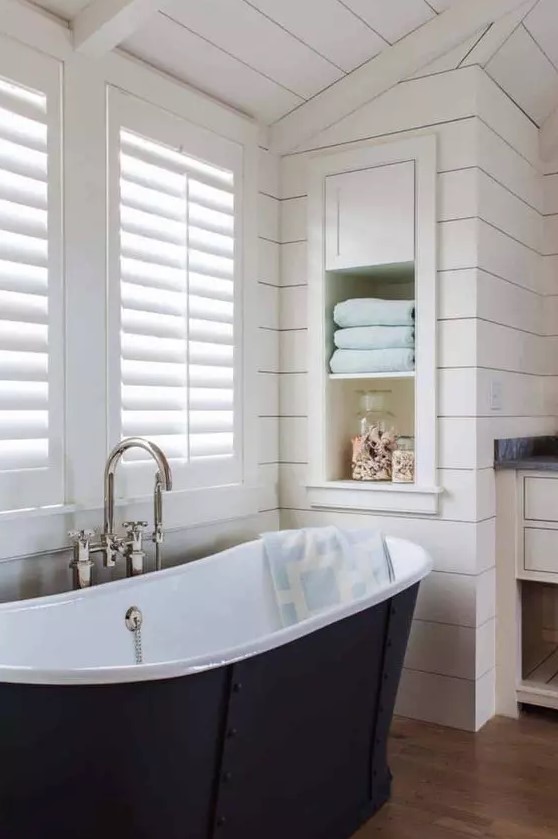 a neutral farmhouse bathroom with planked walls and a ceiling, shutters that match the neutral walls and a navy bathtub