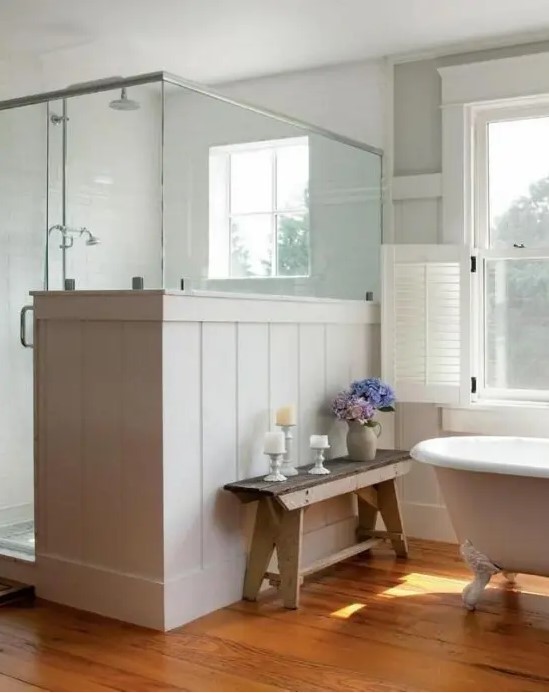 a neutral farmhouse bathroom with planked walls and a shower space, with a pony wall and a vintage bathtub plus a bench