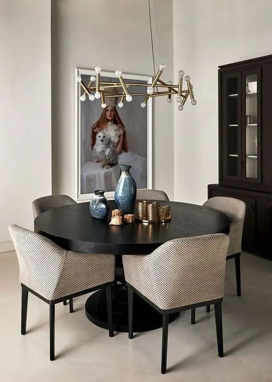 a pretty greige dining room with a dark stained storage unit, a black round table, greige chairs, a gilded chandelier and a large and bold artwork