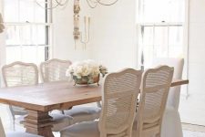 a refined neutral dining room with a stained table, whitewashed chairs with cane backs, a lovely chandelier and much natural light