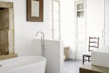 a relaxed neutral bathroom with a wooden beam, an oval tub, a half wall that separates the toilet from the rest of the space