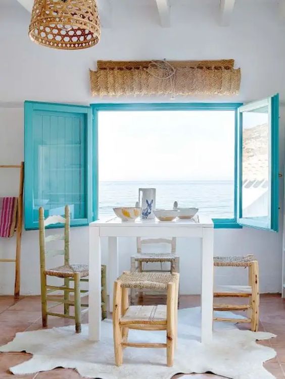 a seaside dining room with a white table and stained chairs, turquoise shutters and a woven pendant lamp