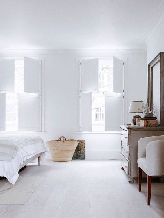 a serene and airy bedroom with a neutral bed and bedding, a whitewashed dresser, a neutral chair, a mirror and white solid shutters
