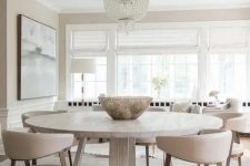 a serene neutral dining room with a round table, neutral chairs, an oversized crystal chandelier and a beautiful artwork