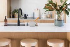 a sleek minimalist blonde wood kitchen with white countertops and a backsplash, black fixtures and some magnolia leaves