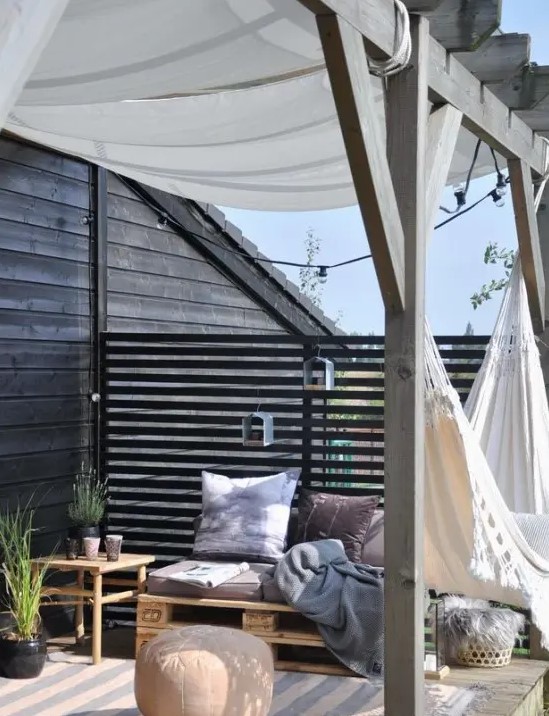 a small Nordic summer terrace with dark walls, pallet and wooden furniture, dusty textiles and a hammock plus ottomans