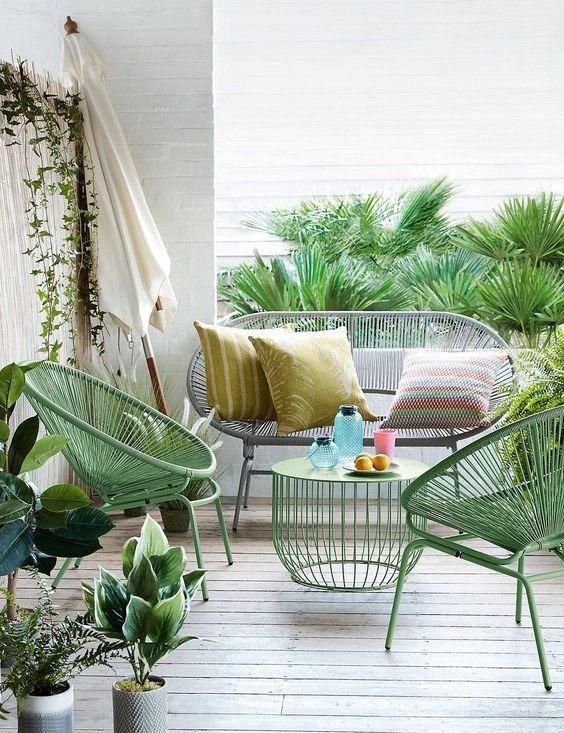 a small and bright terrace with cool mid century modern seating furniture, printed pillows, a lovely green coffee table and lots of greenery around