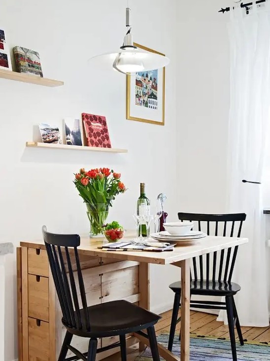 a small and cozy dining space with a folding table, black chairs, open shelves, books and artworks is cozy space