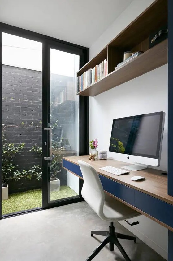 a small and lovely contemporary home office with a floating desk with navy drawers, a wall-mounted bookshelf, a creamy chair and an entrace to a mini garden