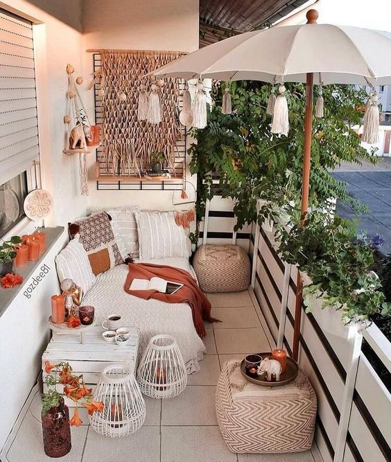 a small and very cozy boho terrace with a sofa and pillows, a white crate side table, an umbrella with tassels, macrame and rust decor