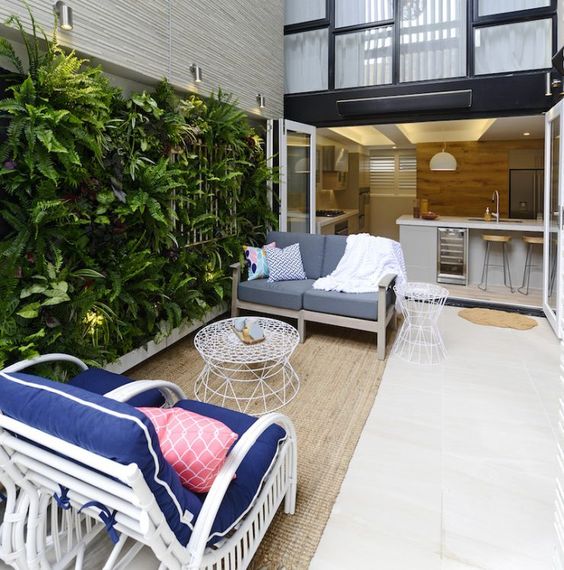 a small and welcoming terrace with a white rattan chair, a living wall, a neutral and grey sofa, a white metal coffee table and spotlights