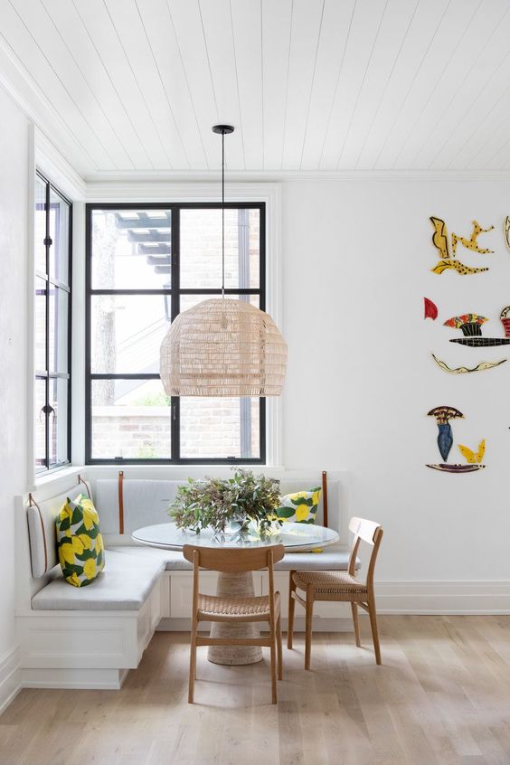 a small bright dining space at the window, with a corner bench, a round table and wooden chairs, a woven pendant lamp