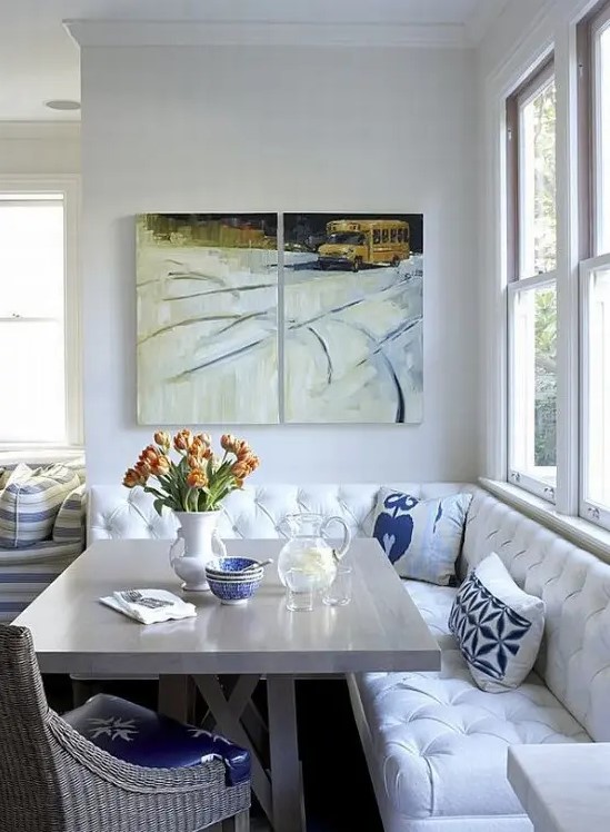 a small dining nook with a white upholstered corner bench, a grey table, a woven chair, some artworks and bold pillows