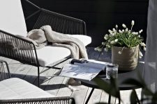 a small modern terrace with metal and wicker chairs, a black table, some simple blooms in a pot and a weathered deck