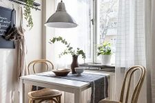 a small vintage dining nook with a white table, light stained chairs, a pendant lamp, greenery and a cute view