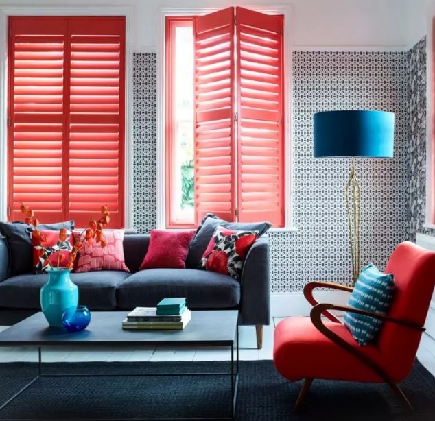 a stylish mid century modern living room with printed wallpaper, a black rug, a grey sofa with red printed pillows, a red chair and matching red shutters to add extra interest