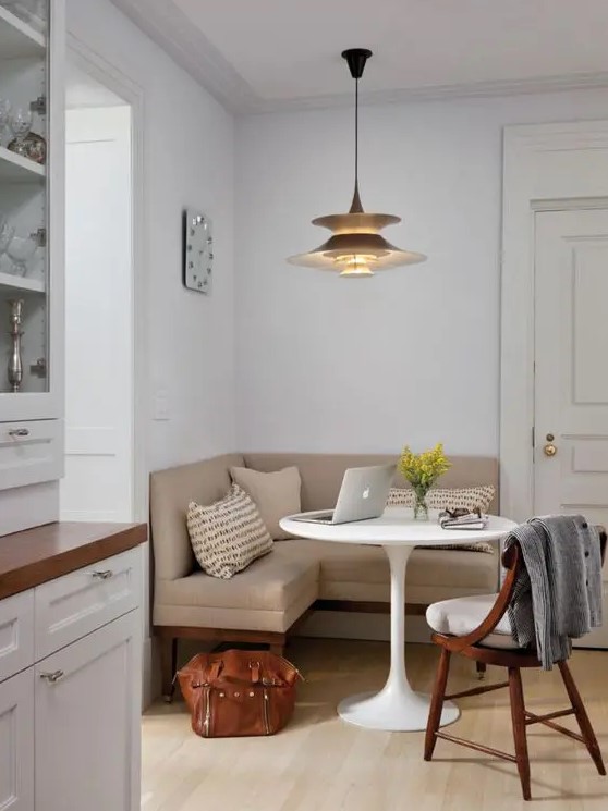 a stylish small dining nook with a tan corner bench, a round table, a white chair, a chic pendant lamp, printed pillows