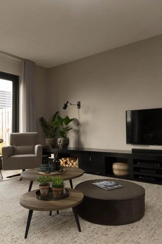 a taupe living room with a taupe chair, a duo of plywood coffee tables, a black TV unit, a brown round ottoman and potted plants