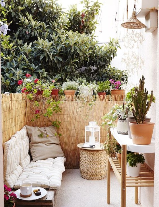a tiny boho terrace with an upholstered bench, a wicker ottoman, a large plant stand and lots of potted plants around