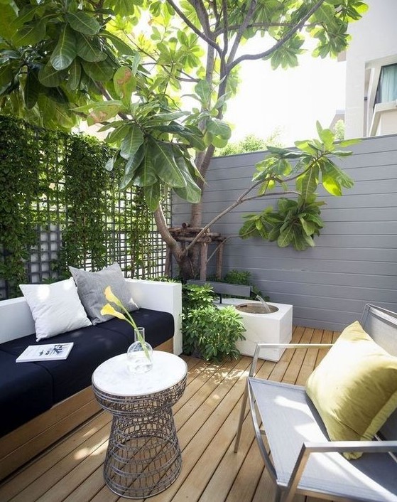 a tiny modern terrace with a wooden deck, a wall with greenery, a built in sofa, a chair and a fountain plus a small table