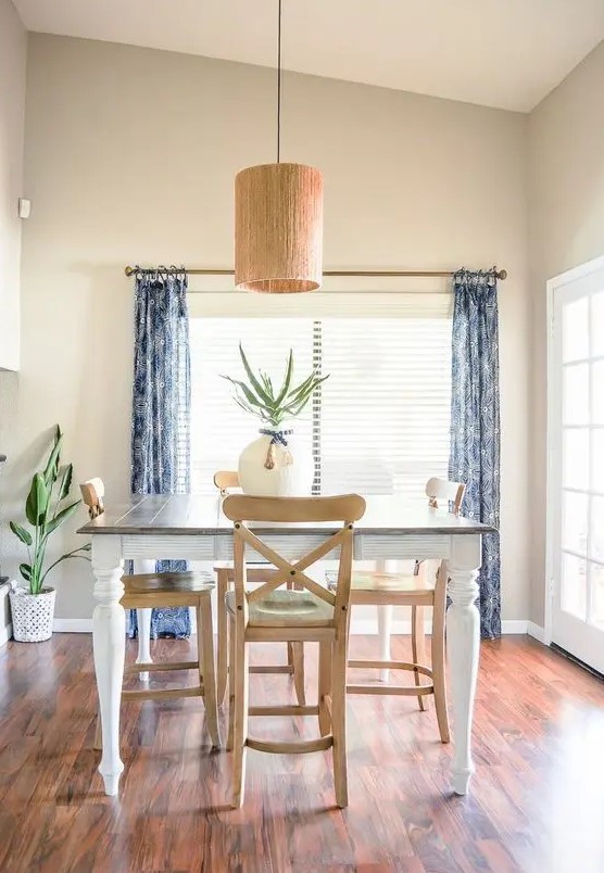 a very simple and relaxed greige dining space with a dining table and stained chairs, blue printed curtains, a pendant lamp