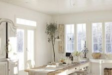 a vintage all-white dining room with extensive glazing, a vintage white table with chairs and benches, a buffet and a console table plus some greenery