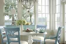 a vintage neutral and pastel dining space with extensive glazing, lake views, a shelf with bottles, a vintage white round table and blue and white chairs plus a large and chic chandelier