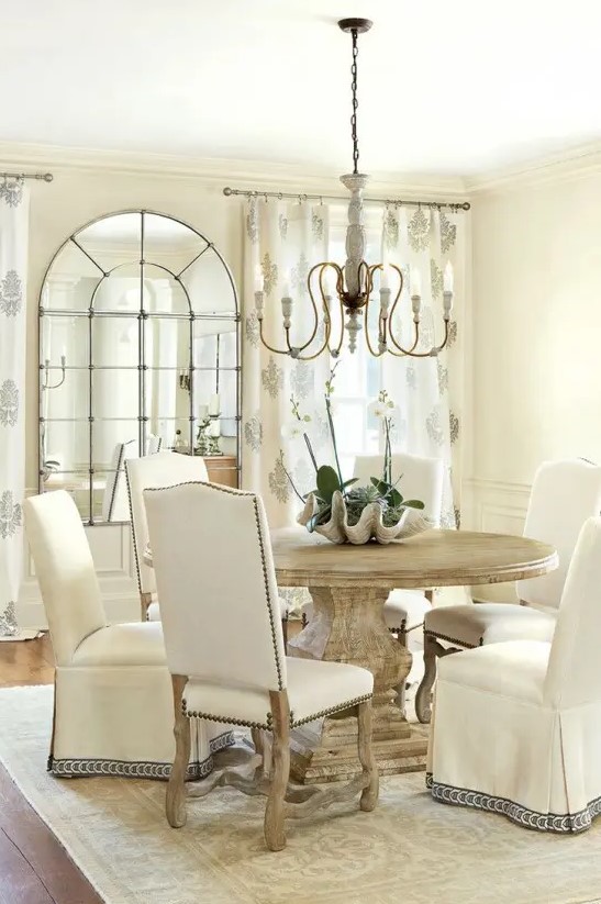 a warm-colored vintage dining room with tan walls, a round stained table and chairs covered with fabric, with a vintage chandelier and a French window