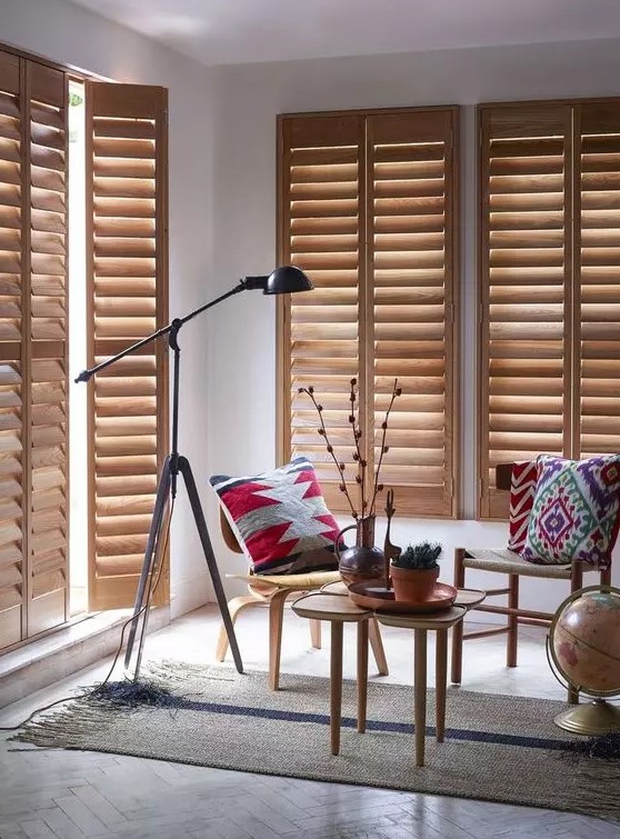 a welcoming living room with stained furniture, bright printed textiles, a black floor lamp, light stained wooden shutters for privacy and blocking excessive sunshine