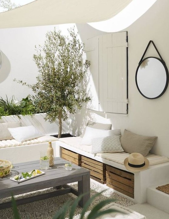 a white Mediterranean terrace with built in benches, greenery and a tree, crates for storage and a small roof