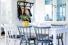 an airy Scandinavian dining space with a neutral cupboard with glass doors, a white vintage dining table, white and blue chairs and a bright vase with blooms