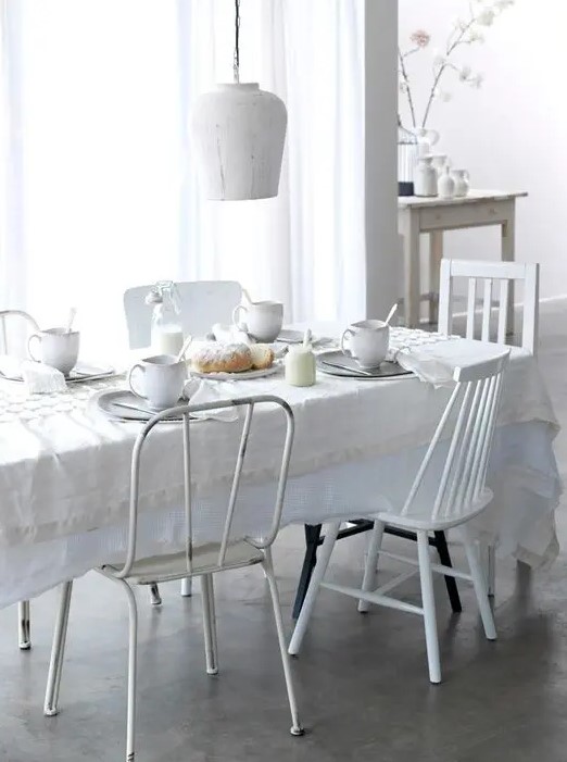 an all white and serene Scandinavian dining room with a table covered with a white tablecloth, mismatching white chairs and a white pendant lamps is super inviting