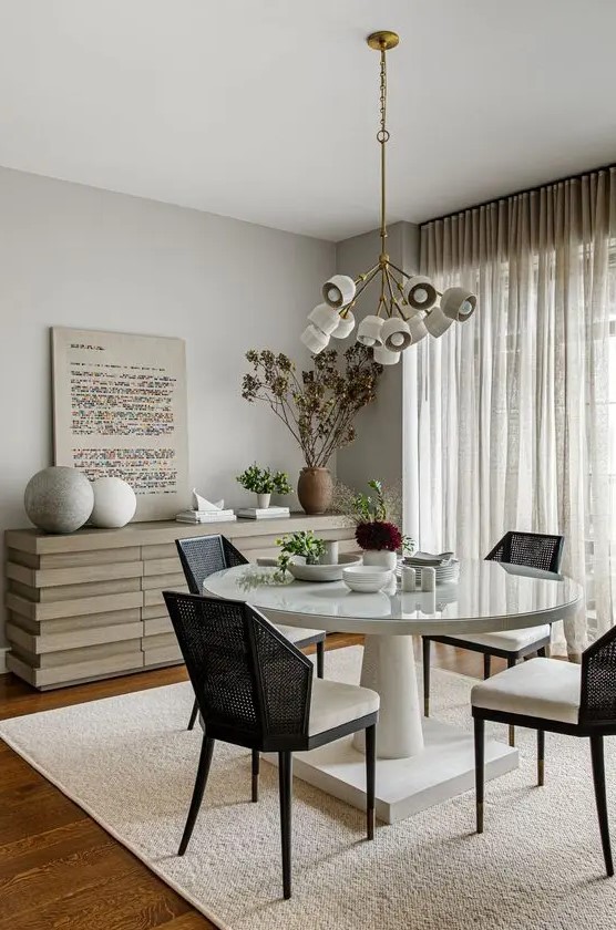 an elegant and cool greige dining room with a catchy credenza, a round table, black chairs, a cool chandelier, some rocks and potted plants