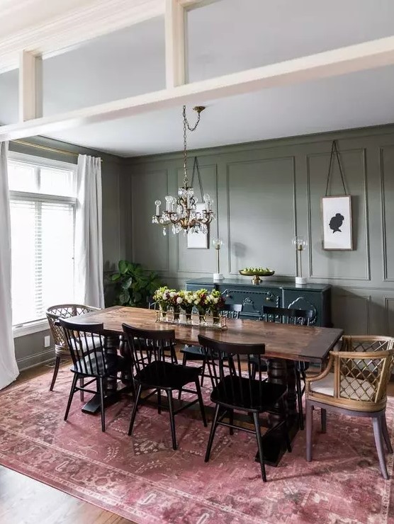 an elegant vintage-inspired dining room with olive green walls, a stained dining table, black wooden and stained rattan chairs, a chic chandelier