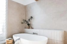 an ethereal greige bathroom with large scale tiles, a wooden beam, a window and a skylight, an oval tub and a wooden side table