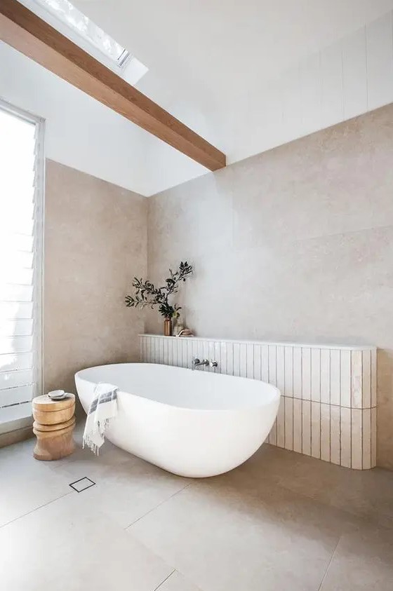 an ethereal greige bathroom with large scale tiles, a wooden beam, a window and a skylight, an oval tub and a wooden side table