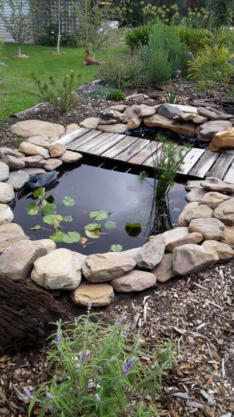 a small natural-looking pond surrounded with rocks, with a wooden bridge over it and cool plants all around