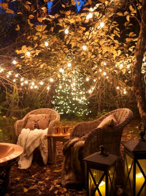 a stylish and cozy backyard with woven chairs, a table, some candle lanterns and string lights over the space