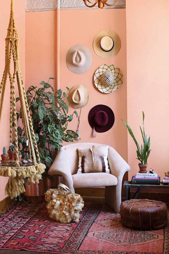 a peachy boho chic space with a burgundy printed rug, a suspended side table, a blush chair, potted plants and hats on the wall