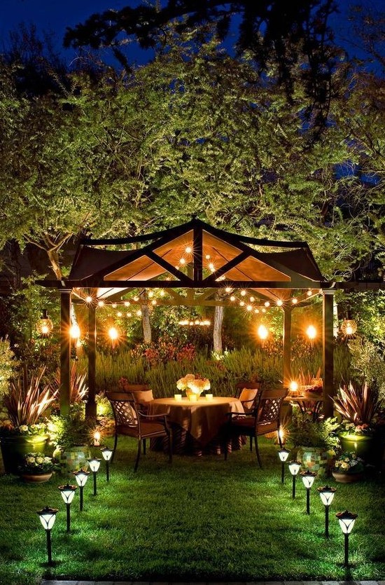 a backyard dining zone with lights over the space and outdoor lamp lining up the path looks very welcoming